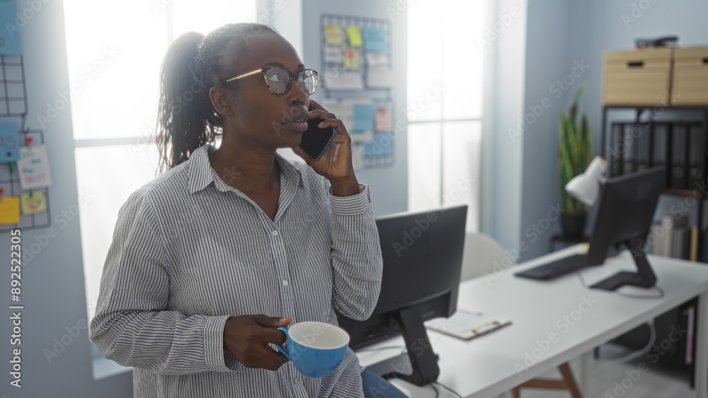 Young african american woman in a workplace holding a coffee cup while talking on the phone, in a modern office setting with computers and a bulletin board Young african american woman in a workplace holding a coffee cup while talking on the phone, in a modern office setting with computers and a bulletin board
