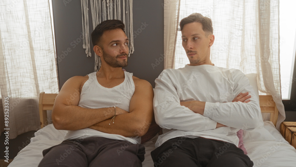 Two men with serious expressions and crossed arms sit on a bed in a cozy bedroom, emphasizing their relationship dynamics and the intimate indoor setting. Two men with serious expressions and crossed arms sit on a bed in a cozy bedroom, emphasizing their relationship dynamics and the intimate indoor setting.