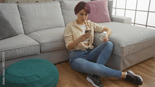 Young woman with short hair sits on the floor of her living room, holding a bottle and looking at her smartphone near a grey couch and teal pouf.