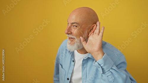 Mature bald man with a grey beard, wearing a blue shirt, holding his hand to his ear while smiling against a yellow background wall.