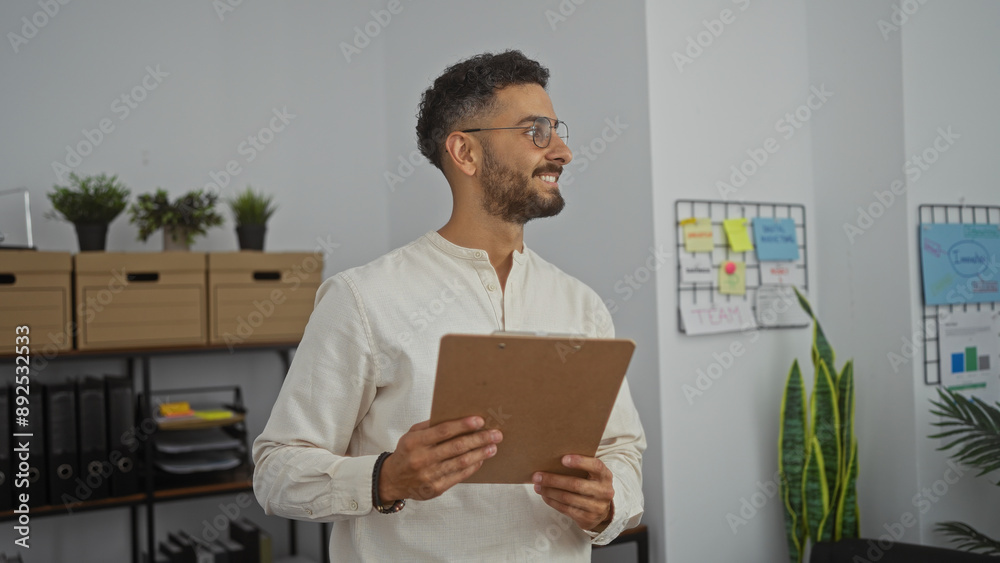 Handsome young hispanic man holding a clipboard while standing in an office workspace with organized files and plants visible in the background. Handsome young hispanic man holding a clipboard while standing in an office workspace with organized files and plants visible in the background.