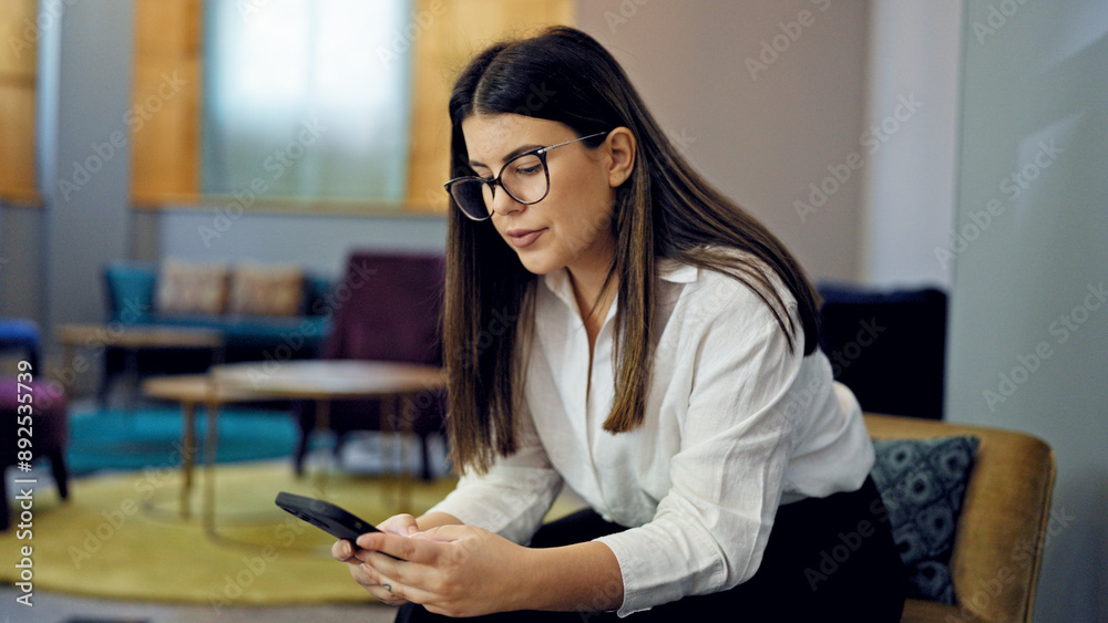 Young beautiful hispanic woman using smartphone sitting on a chair at the office Young beautiful hispanic woman using smartphone sitting on a chair at the office