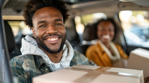 A cheerful man sitting in a car, holding a box, looking directly at the camera, with a smiling woman blurred in the background. This image captures the essence of teamwork and cooperation.