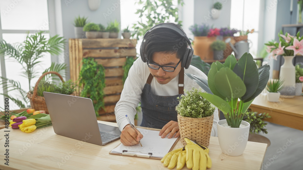 A young asian man with headphones writes notes in a flower shop brimming with greenery and vibrant blooms. A young asian man with headphones writes notes in a flower shop brimming with greenery and vibrant blooms.