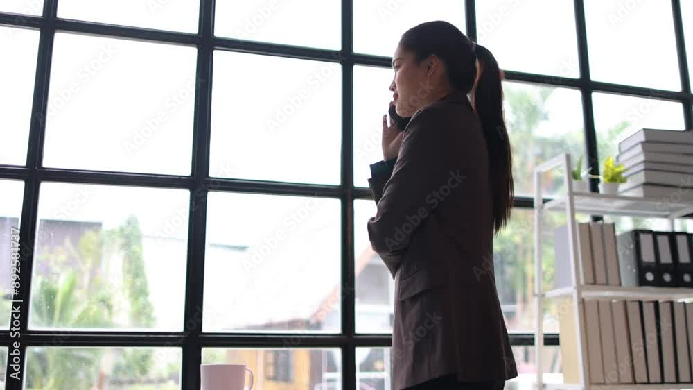 Professional businesswoman on phone by window. Young businesswoman in a professional office environment, having a phone conversation near a bright window, she is smiling happily. Professional businesswoman on phone by window. Young businesswoman in a professional office environment, having a phone conversation near a bright window, she is smiling happily.