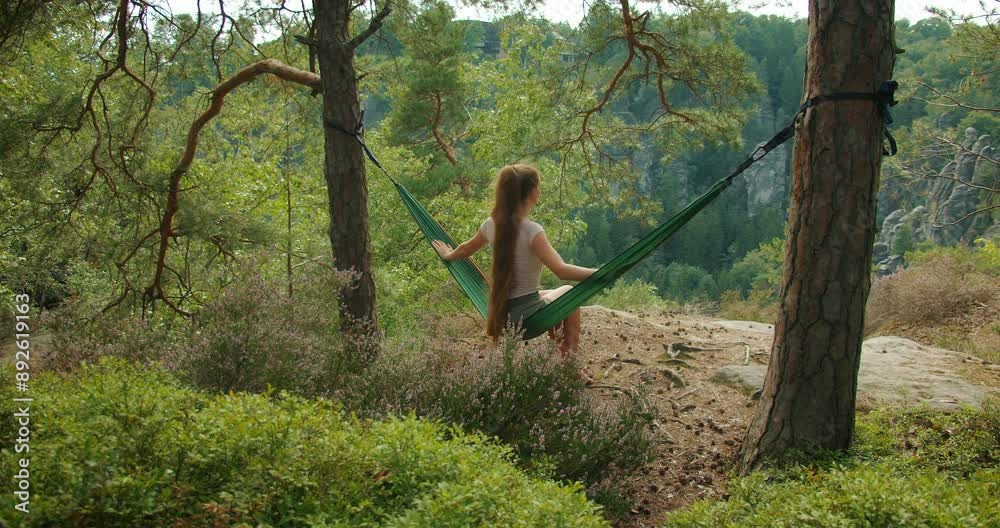 Woman sitting in a hammock in a lush forest, enjoying a peaceful view of the rocky landscape and greenery in summer sunny day