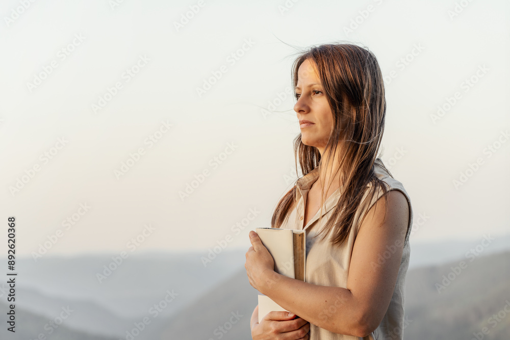 A woman is peacefully standing outside, holding a book in a tranquil natural setting A woman is peacefully standing outside, holding a book in a tranquil natural setting
