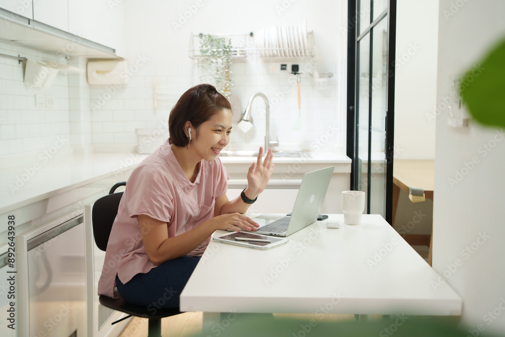 Woman in pink shirt video conferencing on laptop in a modern kitchen, smiling and waving, wearing wireless earphones Woman in pink shirt video conferencing on laptop in a modern kitchen, smiling and waving, wearing wireless earphones