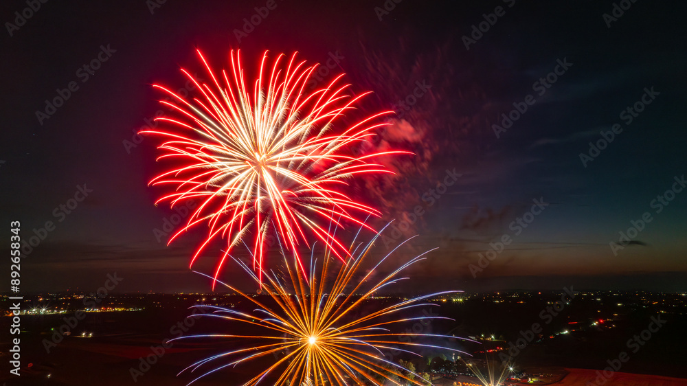 Red And Gold Fireworks Explode Brilliantly In The Night Sky Over A Rural Landscape, Creating Stunning Patterns Of Light Above A Vibrant Fairground, With Distant City Lights On The Horizon. Red And Gold Fireworks Explode Brilliantly In The Night Sky Over A Rural Landscape, Creating Stunning Patterns Of Light Above A Vibrant Fairground, With Distant City Lights On The Horizon.