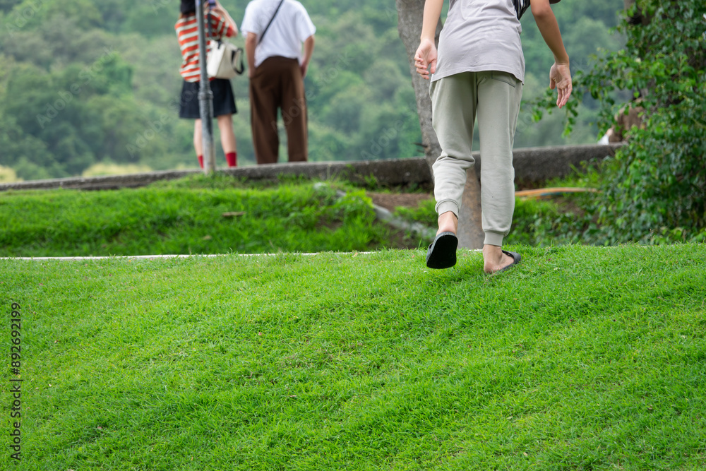 Fototapeta premium A woman is walking on a grassy hill with her feet bare. She is wearing a gray shirt and gray pants. There are other people in the background, but they are not the main focus of the image