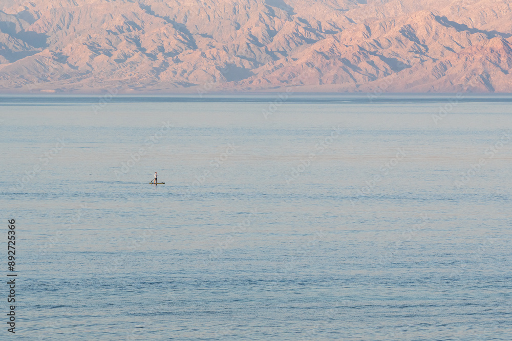 Obraz premium A person on a sap board in Gulf of Aqaba of Red Sea on the background of Saudi Arabia's mountains on sunset. Dahab, Egypt.