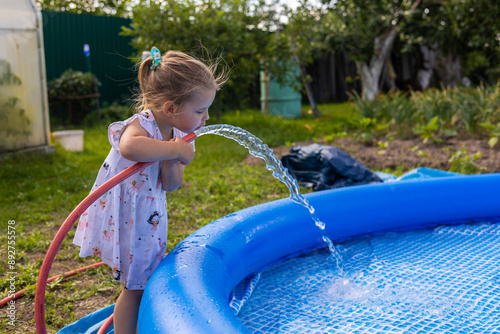 Cute little child pours water into the pool from a hose. A little girl bends down and drinks cool water from a hose as it fills the pool. In summer, the child fills the pool with water.