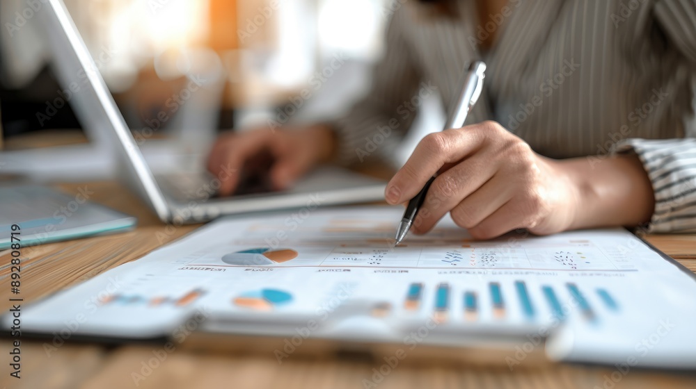 A person reviews charts and graphs on a document with a laptop in the background, symbolizing data analysis, business planning, or financial review. A person reviews charts and graphs on a document with a laptop in the background, symbolizing data analysis, business planning, or financial review.
