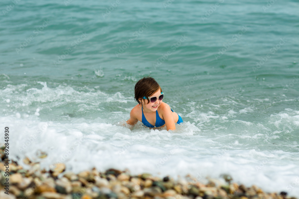 A small, beautiful girl in a blue swimsuit and sunglasses, swimming and having fun on the shore, in the water A small, beautiful girl in a blue swimsuit and sunglasses, swimming and having fun on the shore, in the water
