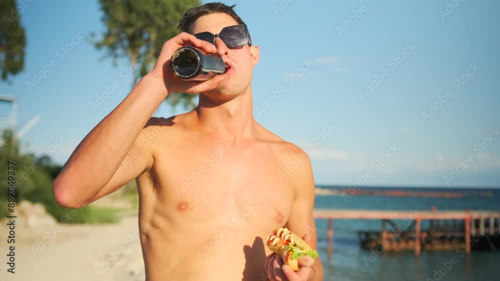 Attractive male tourist eating a hot dog while drinking a bottle of beer while walking along the beach next to the pier and yacht. Attractive male tourist eating a hot dog while drinking a bottle of beer while walking along the beach next to the pier and yacht.