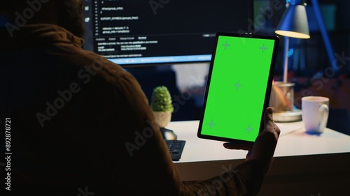 Software developer programming in home office, crosschecking lines of code between green screen tablet and PC screen. IT admin holding mockup portable device while coding on computer photo