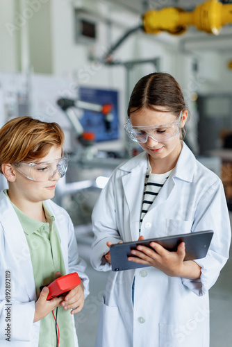 Two classmates standing in robotic laboratory, wearing lab coats and safety eyeglasses. After-school robotics club. Children learning robotics in Elementary school. photo