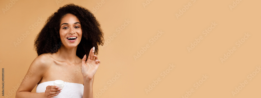 Naklejka premium Skin care concept. Pretty african american lady applying moisturizing face cream, posing wrapped in towel and smiling at camera, standing over brown background, copy space