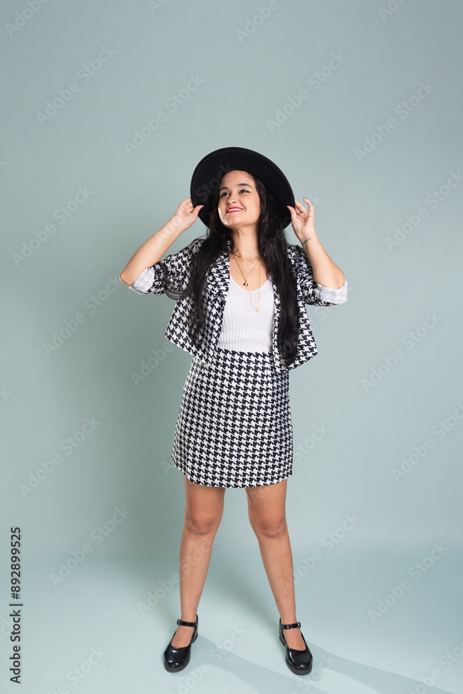 Young beautiful woman standing in studio posing for photo. Wearing and holding a black hat. Calm woman at ease wearing black and white clothing. Young beautiful woman standing in studio posing for photo. Wearing and holding a black hat. Calm woman at ease wearing black and white clothing.