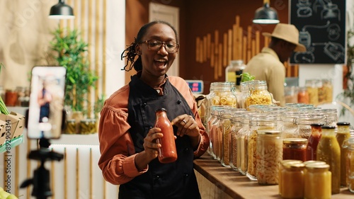 African american vendor promoting homemade sauces and honey, creating online video advertisement to present eco friendly products at zero waste supermarket. Woman recording promotional ad. Camera 1. photo