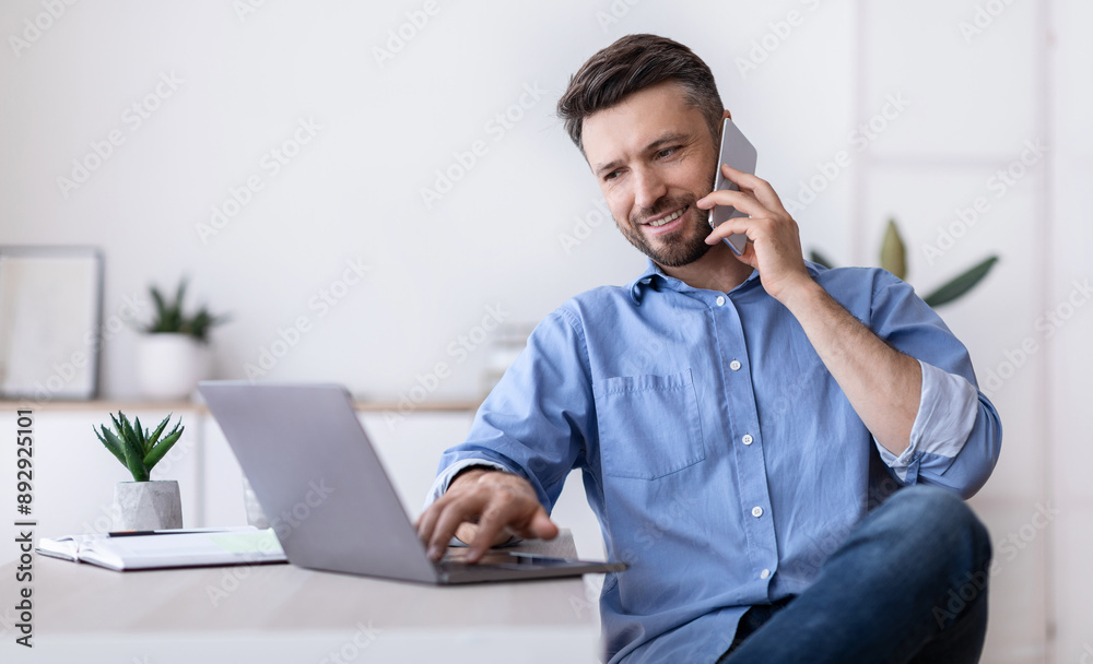 Cheerful Young Man Working Remotely From Home Office With Cellphone And Laptop, Sitting At Desk In Room, Copy Space Cheerful Young Man Working Remotely From Home Office With Cellphone And Laptop, Sitting At Desk In Room, Copy Space