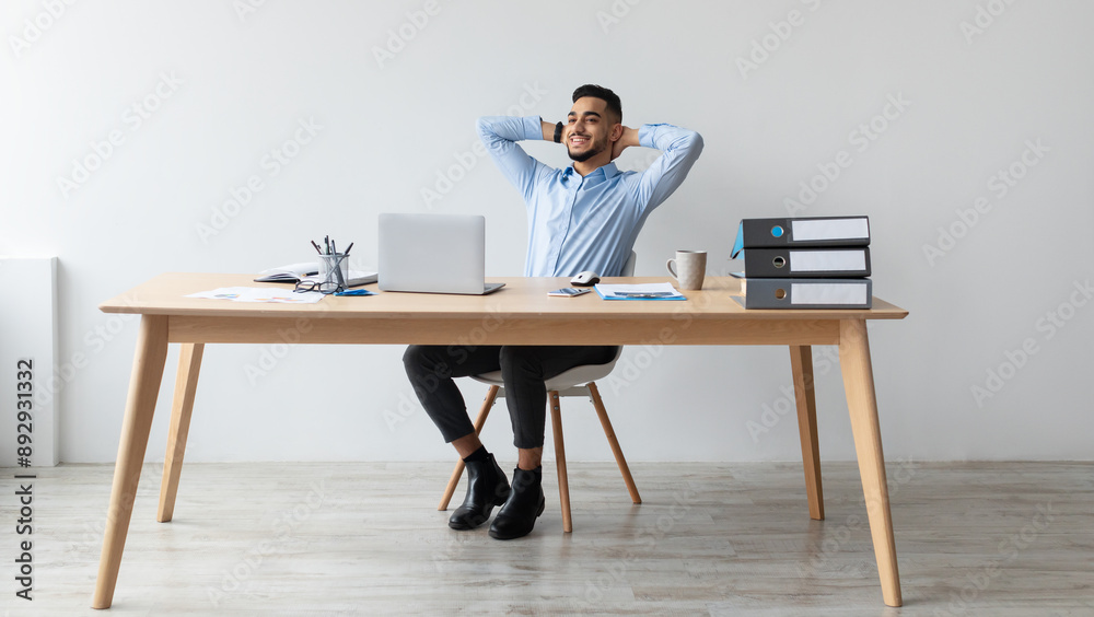Taking Break. Smiling young Arab man relaxing on chair sitting at table and resting, using pc laptop, happy millennial male leaning back at workplace, enjoying his job, feeling pleased and satisfied Taking Break. Smiling young Arab man relaxing on chair sitting at table and resting, using pc laptop, happy millennial male leaning back at workplace, enjoying his job, feeling pleased and satisfied