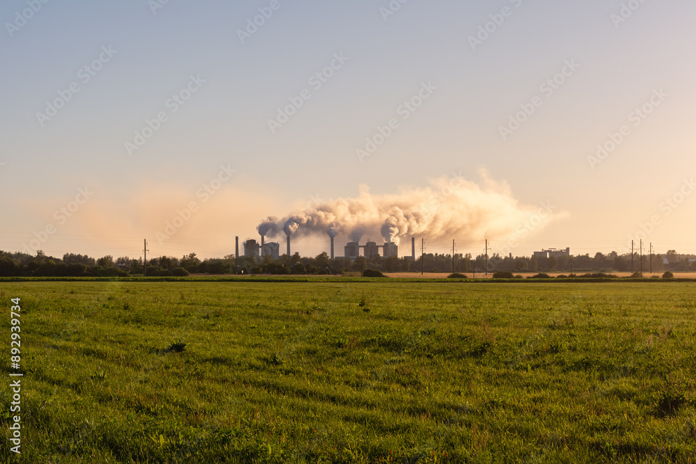 Fototapeta premium A grass field, on the horizon there is a working chemical plant, there are many clouds of smoke from the factory chimneys, illuminated by sunlight. Ecological concept image in summer during sunset