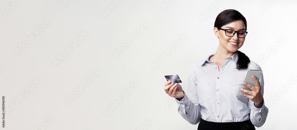 A smiling woman in a white button-down shirt holds a credit card in one hand and a smartphone in the other, looking down at the phone screen. A smiling woman in a white button-down shirt holds a credit card in one hand and a smartphone in the other, looking down at the phone screen.