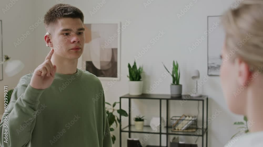 Medium over-shoulder shot of Caucasian male teenager with cochlear implant standing in classroom with female teacher, reciting sign language alphabet letters in ASL, and teacher nodding