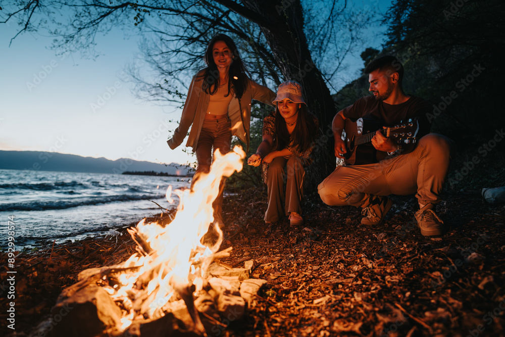 Fototapeta premium Group of three friends gathered around a warm campfire on the beach, enjoying live guitar music under an evening sky.