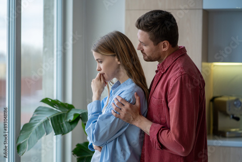 Supportive husband hugging his upset, depressive wife from behind at window in bright apartment. Loss and sorrow lead to depression, grief in family relationship when love and support needed. 
