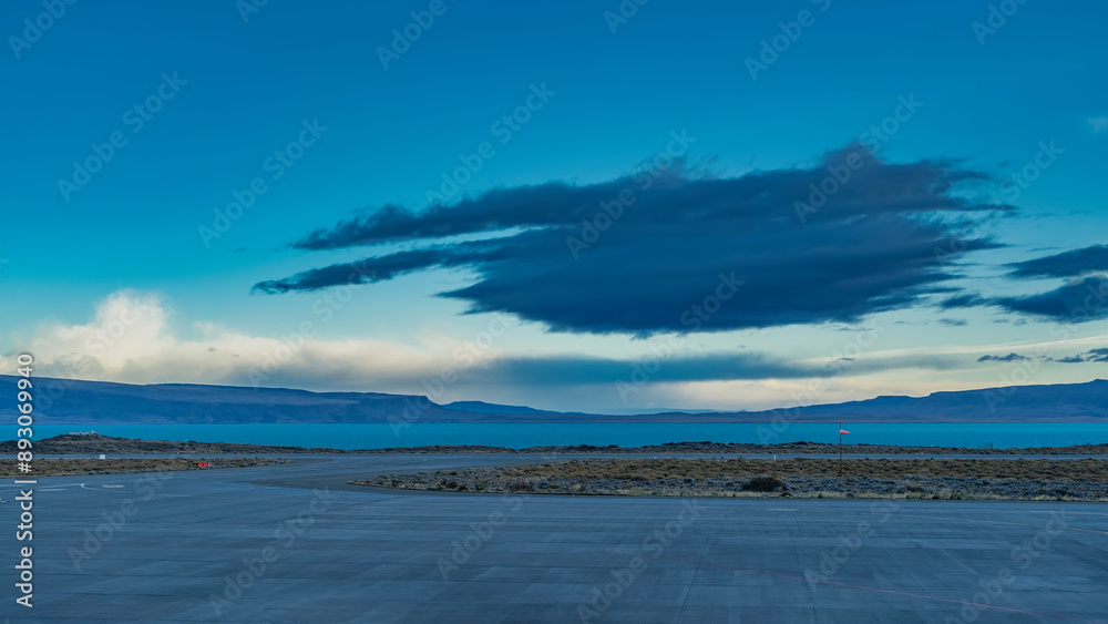 Fototapeta premium The runway of the airfield. The weather vane flutters in the wind. Low-growing grassy vegetation along the road. Turquoise glacial lake in the distance.Mountains against the sky and clouds.El Calafate