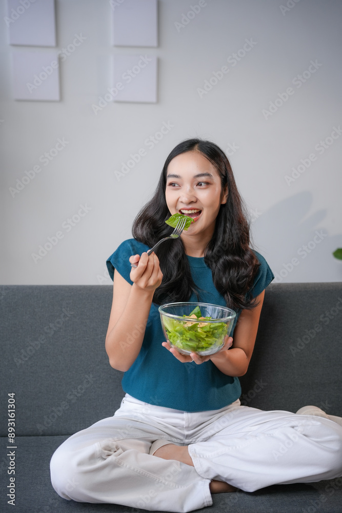 Young woman happily eating a fresh salad on the sofa at home, promoting healthy living and self care with a bright smile, embodying vitality and wellbeing