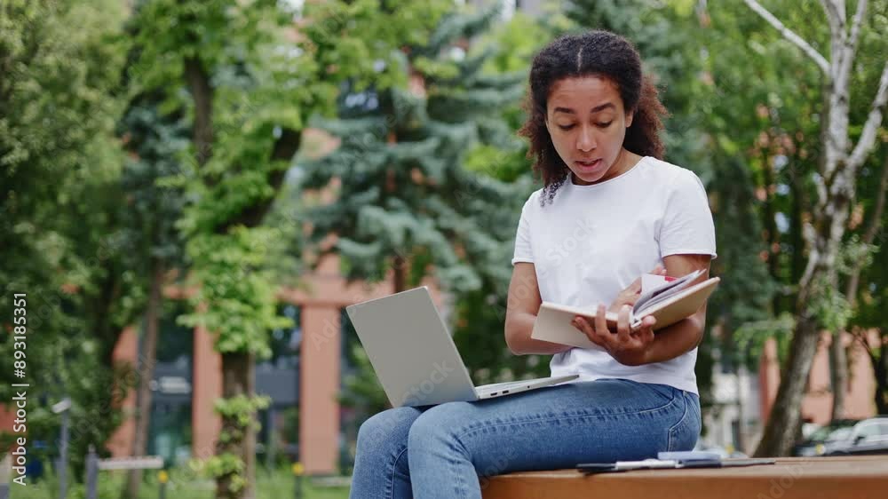 Side view of vibrant african american woman holding personal diary during video communication on wireless laptop outdoors. Concentrated lady engaging in online conversation while sitting outside. Side view of vibrant african american woman holding personal diary during video communication on wireless laptop outdoors. Concentrated lady engaging in online conversation while sitting outside.