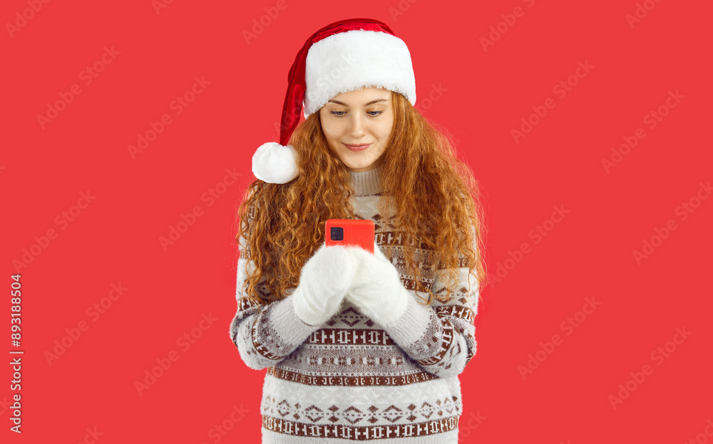 Curly pretty redhead Christmas girl in Santa hat, mittens and knitted sweater with ornament pattern is looking at smartphone on red background. Reading New Year's greetings. Winter holidays concept. Curly pretty redhead Christmas girl in Santa hat, mittens and knitted sweater with ornament pattern is looking at smartphone on red background. Reading New Year's greetings. Winter holidays concept.