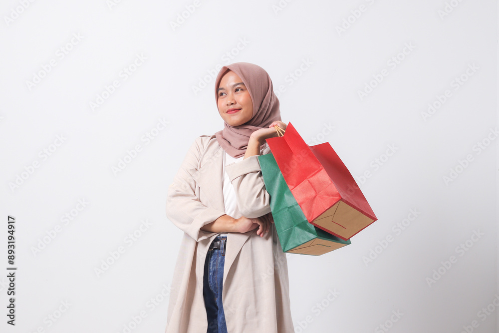 Portrait of cheerful Asian hijab woman in casual suit holding and showing colorful paper shopping bags. Shopaholic girl and discount buying concept. Isolated image on white background Portrait of cheerful Asian hijab woman in casual suit holding and showing colorful paper shopping bags. Shopaholic girl and discount buying concept. Isolated image on white background
