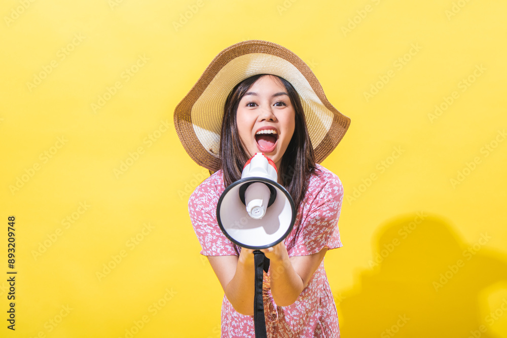 Fototapeta premium Asian woman wearing a straw hat and a pink floral dress, holding a megaphone with a shouting expression against a yellow background. The lighting style is bright and even.