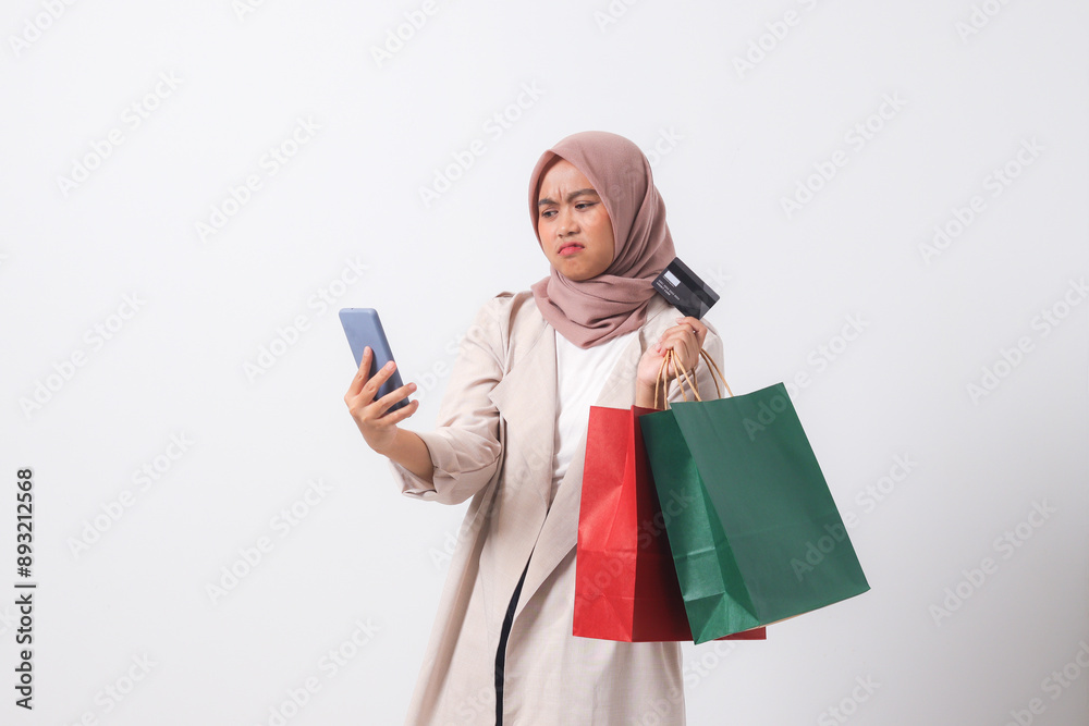 Portrait of excited Asian hijab woman in casual suit holding credit card and shopping bags while using mobile phone. Shopaholic girl and discount buying concept. Isolated image on white background Portrait of excited Asian hijab woman in casual suit holding credit card and shopping bags while using mobile phone. Shopaholic girl and discount buying concept. Isolated image on white background