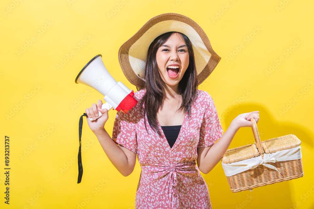 Indonesian Asian woman wearing a straw hat and pink floral dress, holding a megaphone and a wicker basket, smiling and looking at the camera, against a yellow background. bright and even light Indonesian Asian woman wearing a straw hat and pink floral dress, holding a megaphone and a wicker basket, smiling and looking at the camera, against a yellow background. bright and even light
