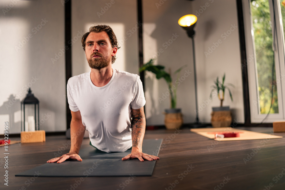 Fototapeta premium A man practicing yoga indoors, performing the upward-facing dog pose on a black yoga mat. The room has wooden floors and other yoga mats and props are visible in the background.