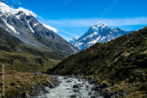 A river flows through Hooker Valley, flanked by lush green hills and snow-capped mountains. The clear blue sky enhances the picturesque alpine scenery. The natural beauty of the valley is captivating