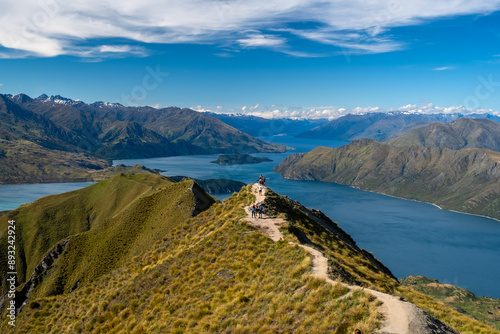 A group of hikers stands on a mountain peak, enjoying expansive views of a tranquil lake and distant mountain ranges. The clear sky and rugged terrain create an atmosphere of adventure and exploratio photo