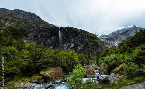 A panoramic view of a river flowing through a dense forest, with lush greenery and rugged mountain backdrop, creating a dynamic natural scene.