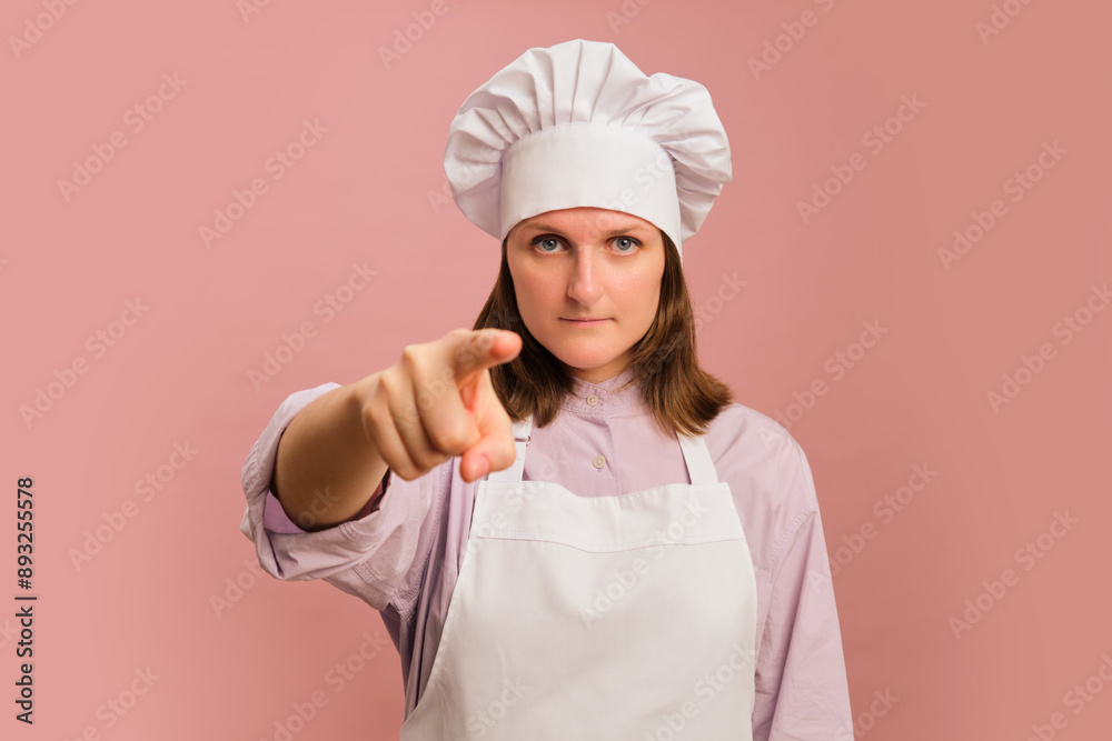 Woman chef pointing finger forward on studio pink background. Portrait of a female person in chef's clothing