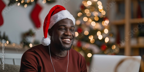 A cheerful man in a Santa hat poses indoors with a laptop, spreading holiday joy.