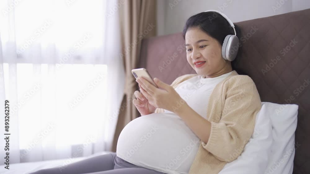 A portrait of happy Asian pregnant woman using online mobile phone technology device with screen in bedroom at home. Having a baby. Family people lifestyle. Mom love. A portrait of happy Asian pregnant woman using online mobile phone technology device with screen in bedroom at home. Having a baby. Family people lifestyle. Mom love.