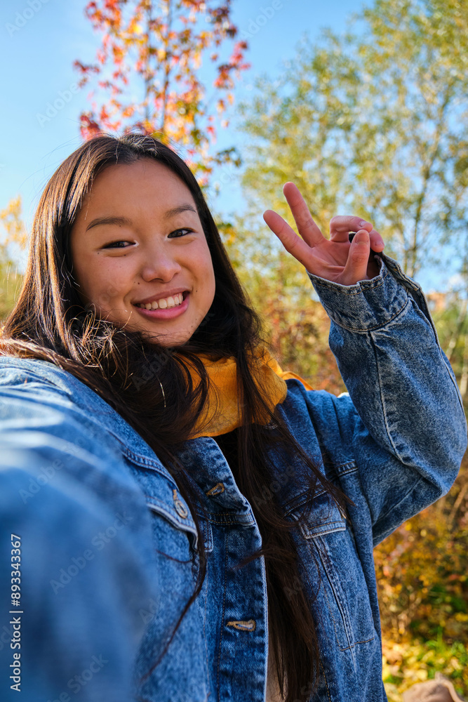 Young Asian woman smiling and taking a selfie with V sign in a park in autumn.