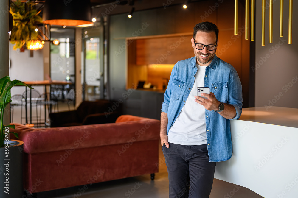 Smiling male freelancer with hand in pocket texting over mobile phone while standing by table in office