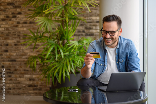 Happy successful businessman making online payment with laptop and credit card on table in office
