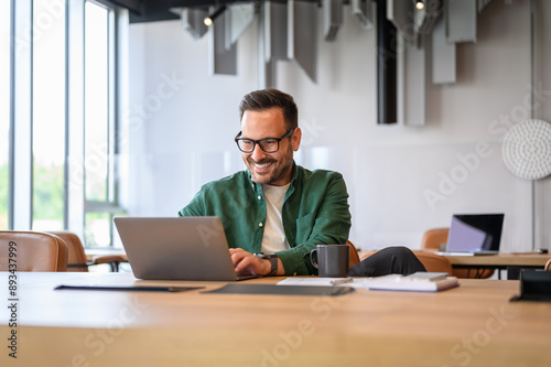 Confident handsome programmer in eyeglasses developing software over laptop at desk in modern office